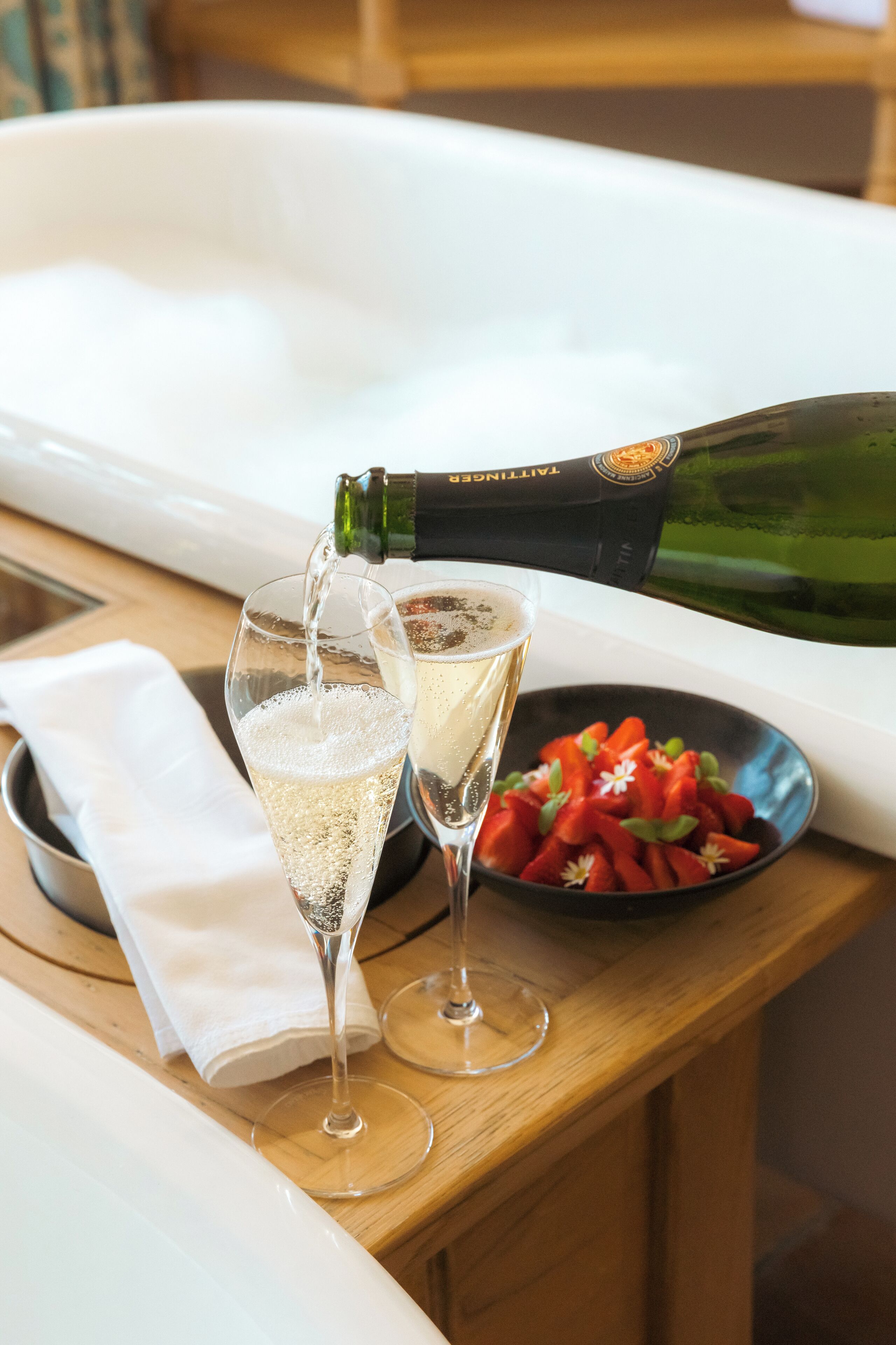A wooden table with two champagne flutes being filled, a bowl of sliced strawberries, and a bottle of champagne.