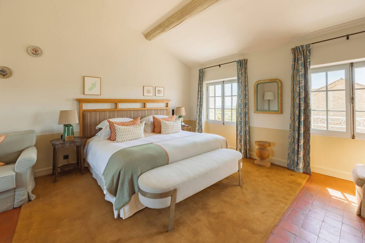 Een slaapkamer met een groot bed, een nachtkastje, een lamp, een bankje en een raam met gordijnen.