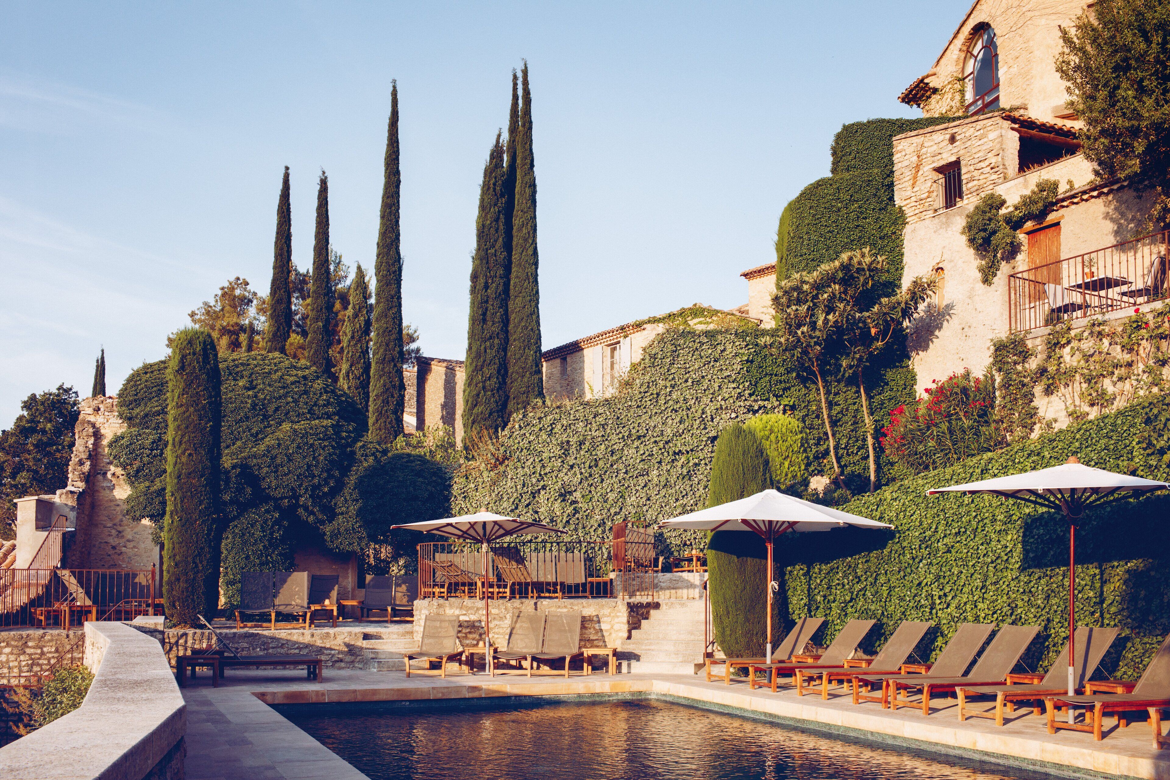 A poolside area with lounge chairs and umbrellas, surrounded by tall cypress trees and a stone building with ivy.