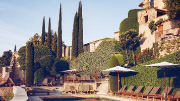 A poolside area with lounge chairs and umbrellas, surrounded by tall cypress trees and a stone building with ivy.