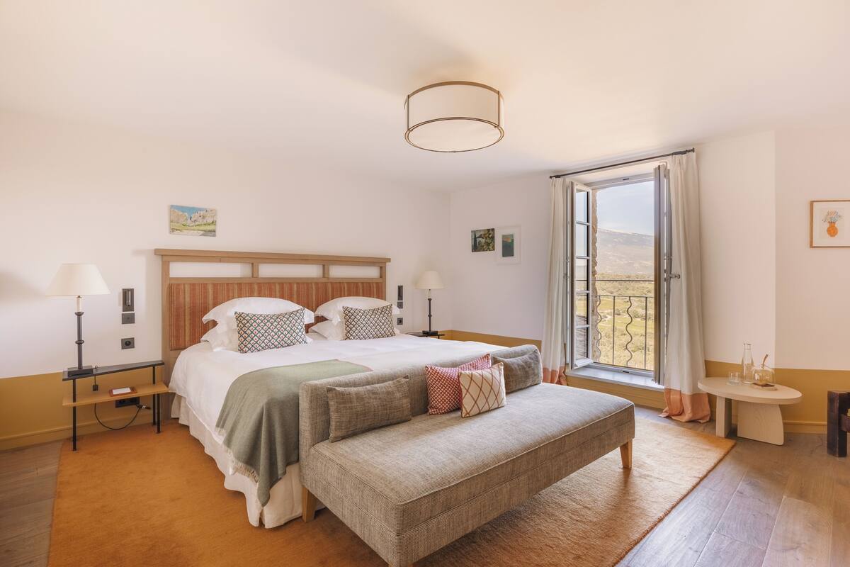 Een slaapkamer met een bed, nachtkastjes, een bankje, een raam met gordijnen en uitzicht op de omgeving.