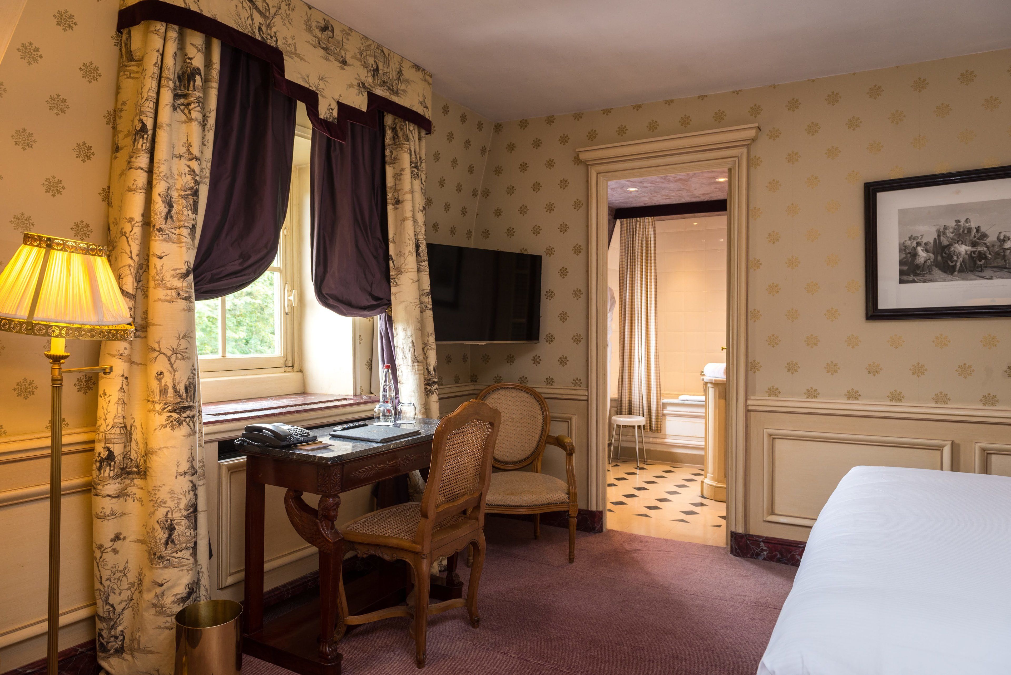 A hotel room with a desk, two chairs, a bed, a television, and a framed picture on the wall.