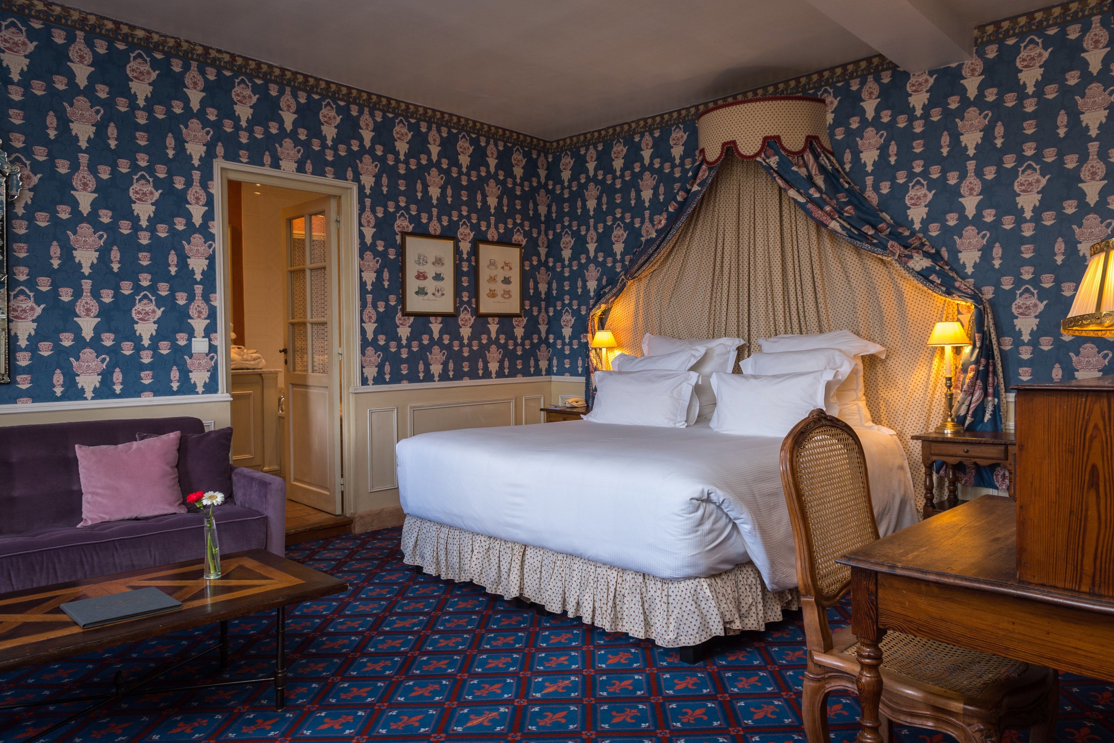 A bedroom with a four-poster bed, a wooden nightstand, a purple sofa, and a patterned carpet.