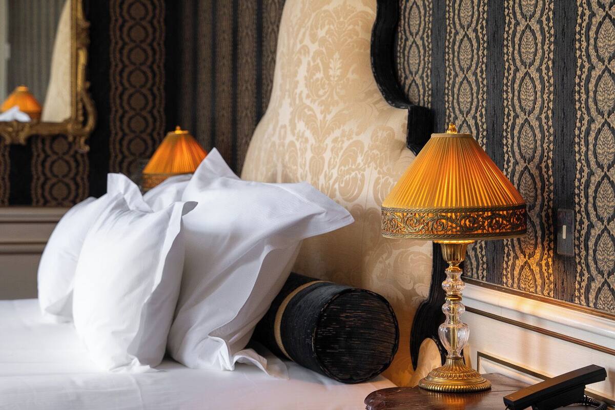 Un lit bien fait, avec du linge de lit blanc, une lampe décorative et un miroir posé sur une commode.