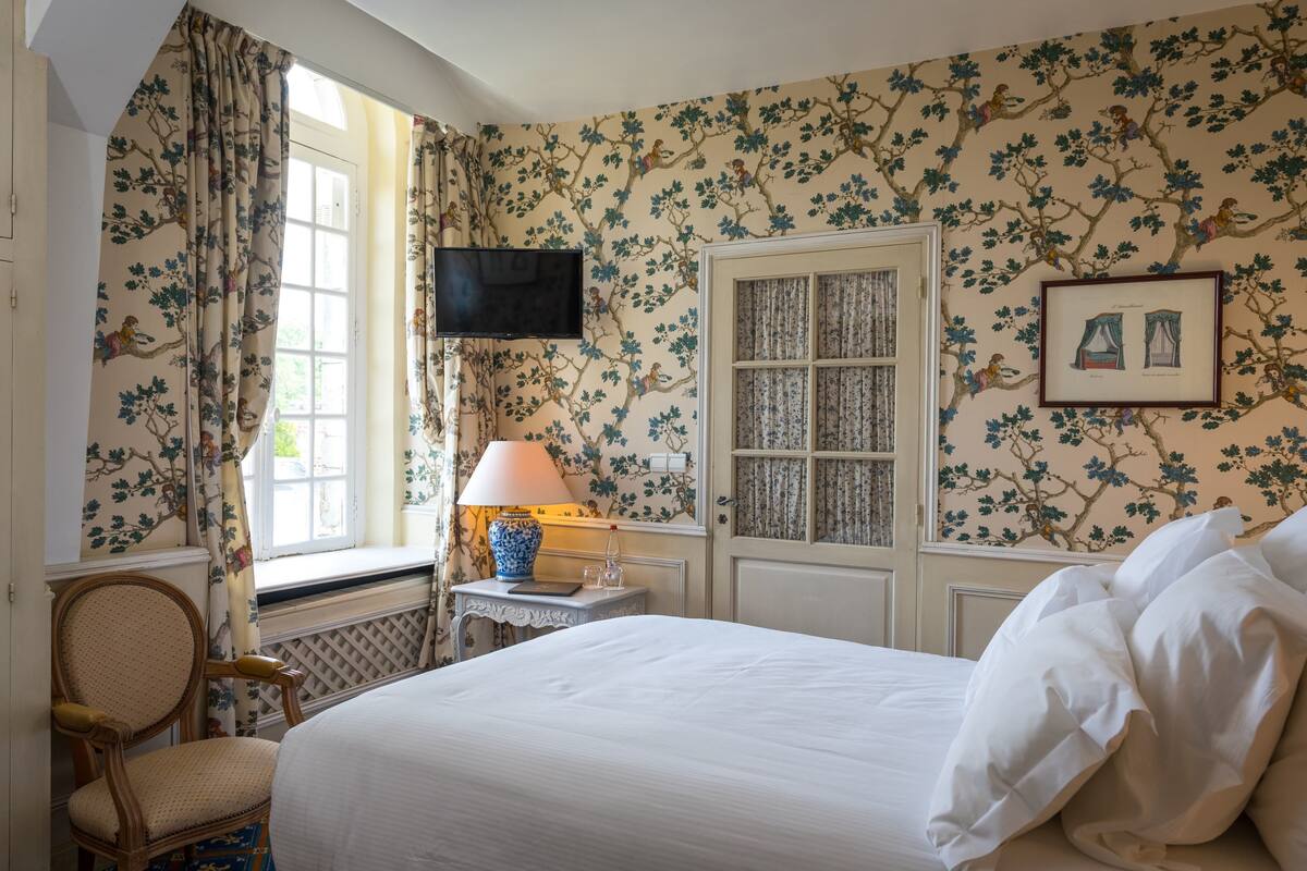 Une chambre d’hôtel avec un lit, une chaise, une table avec une lampe, un téléviseur et une fenêtre avec des rideaux.