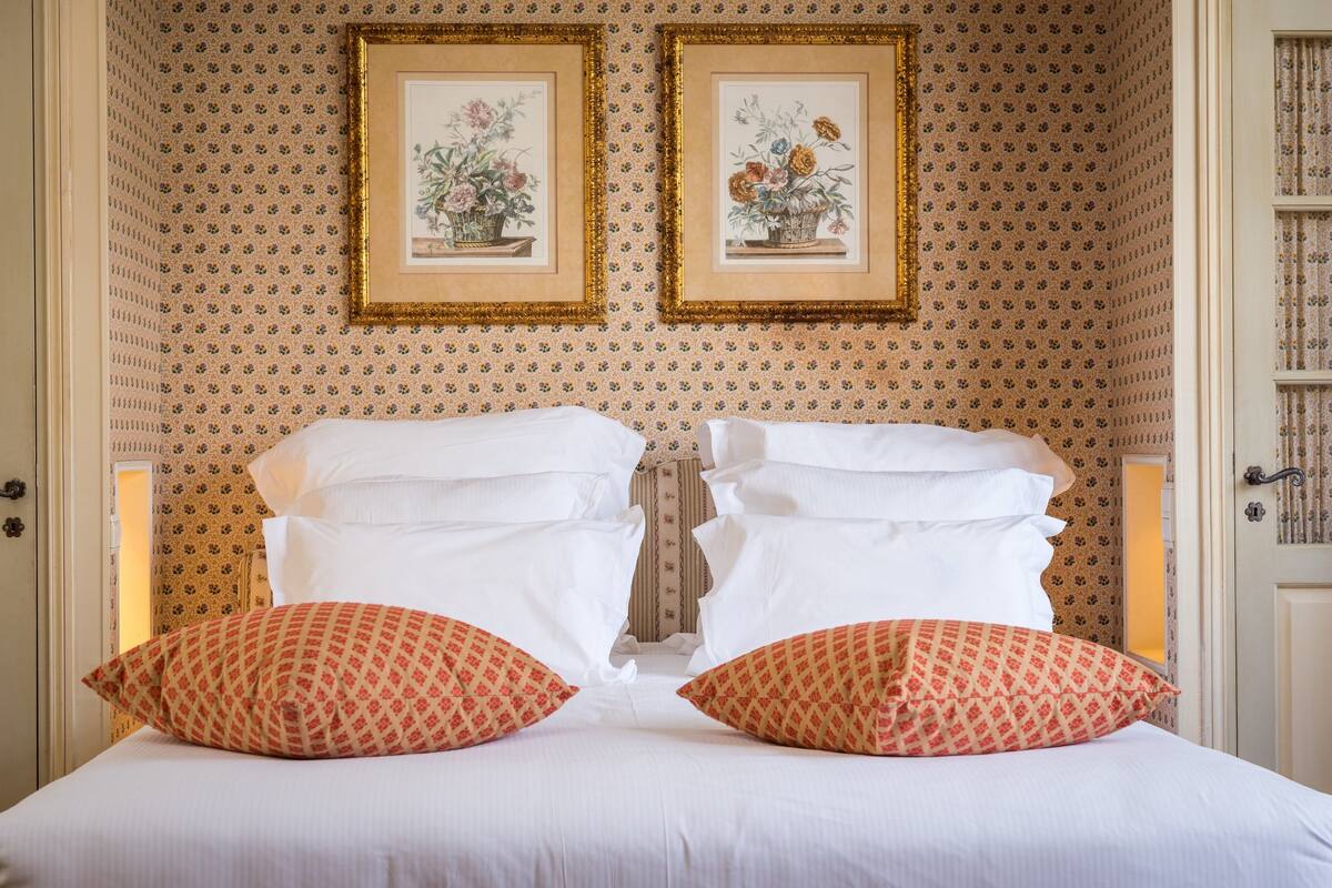 Un lit bien fait, avec du linge de lit blanc et deux coussins à motifs, une œuvre d’art florale encadrée au mur et un papier peint à motifs.