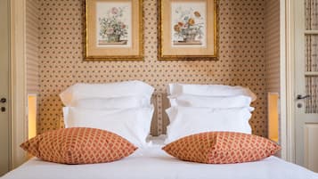 A neatly made bed with white linens and two patterned pillows, framed floral artwork on the wall, and a patterned wallpaper.
