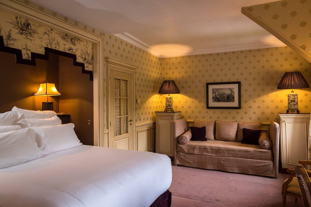 Une chambre d’hôtel avec un grand lit, un canapé, deux lampes, un tableau encadré au mur et un papier peint à motifs.