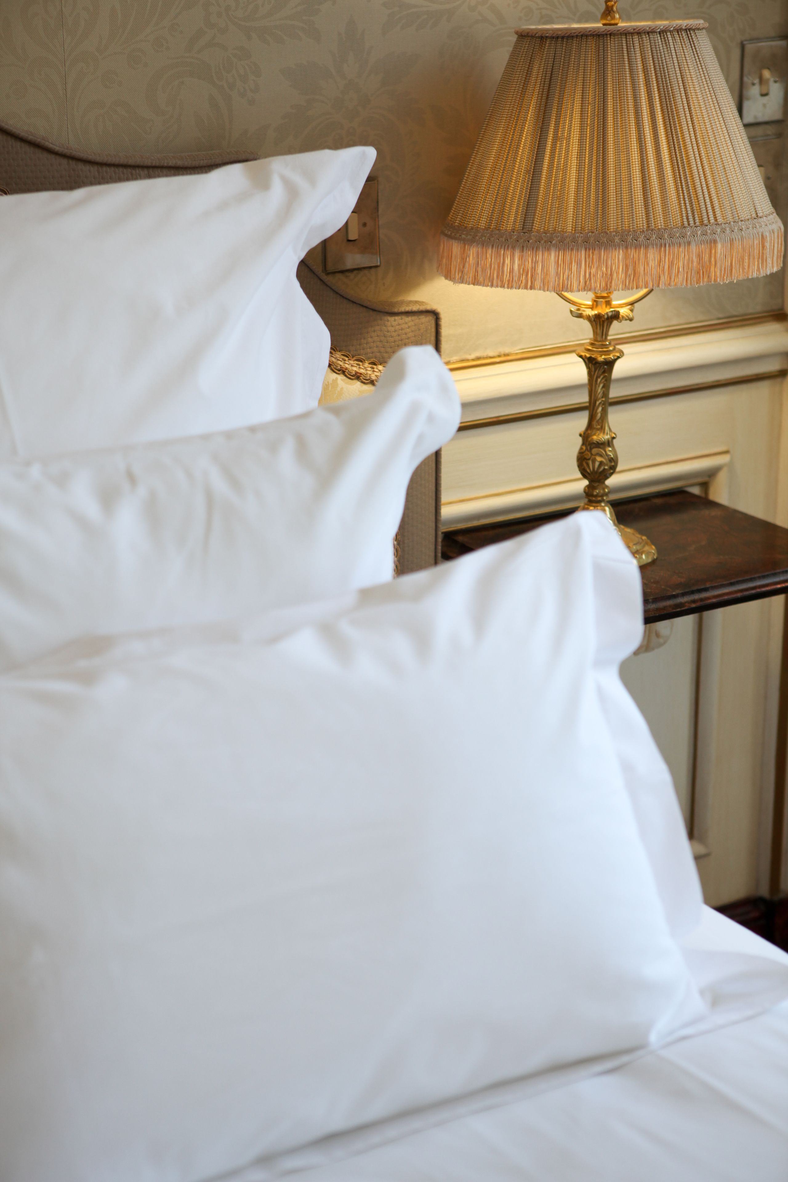 Un lit bien fait, avec du linge de lit blanc et une lampe à abat-jour doré posée sur une table de chevet en bois.