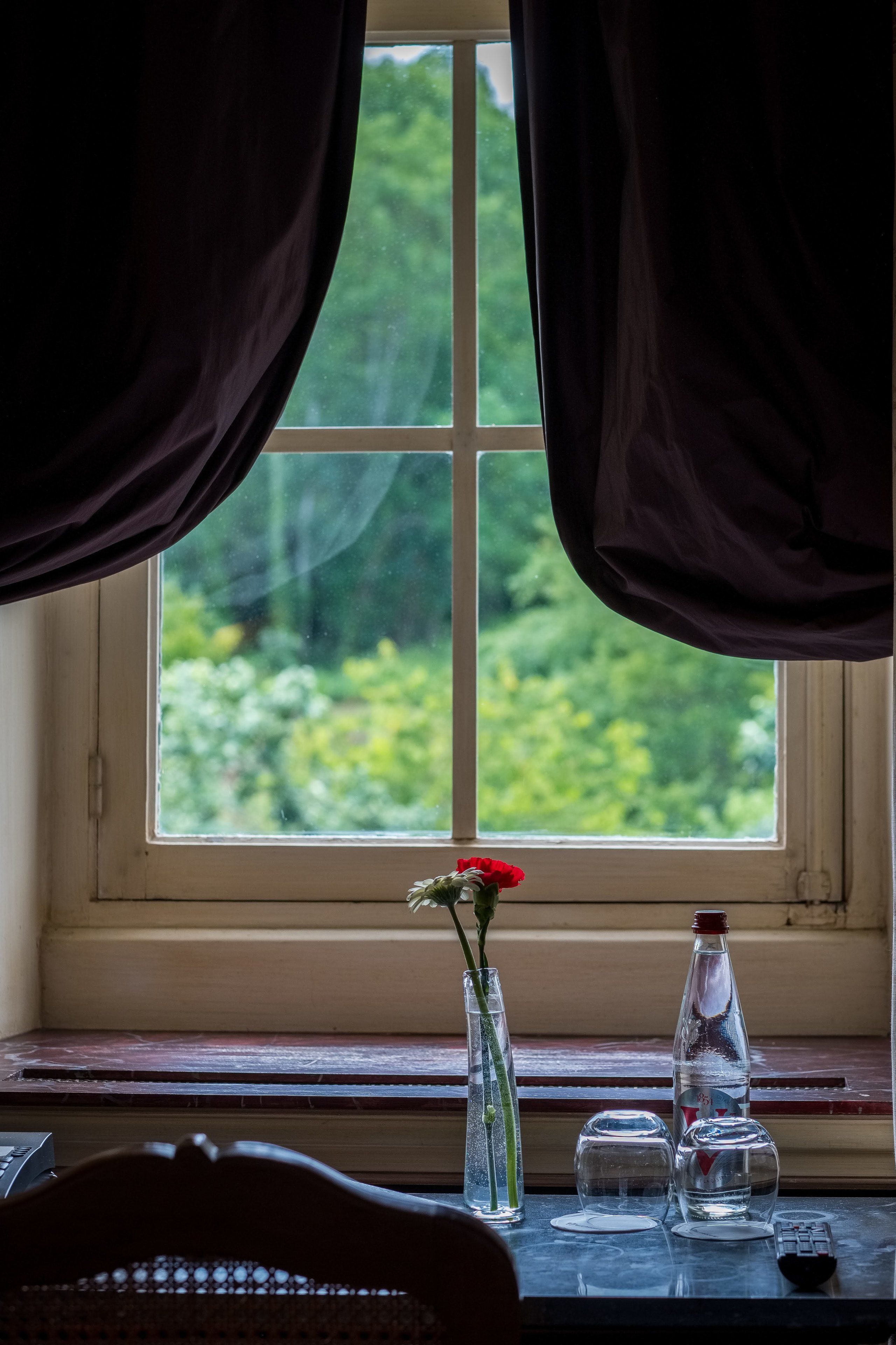 Une fenêtre avec des rideaux, un vase contenant une fleur rouge et une bouteille posée sur une table.