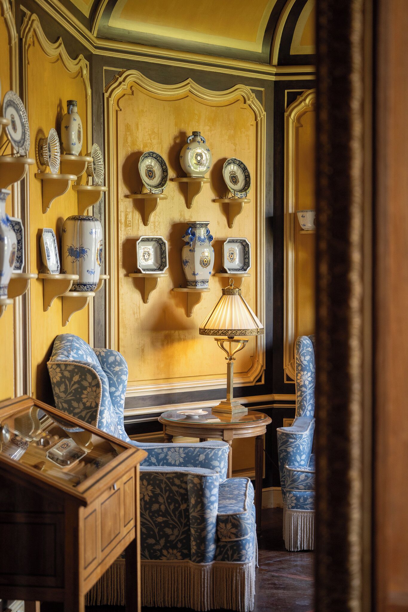 A room with a wall-mounted shelf displaying various ceramic items, a table with a lamp, and patterned blue upholstered chairs.
