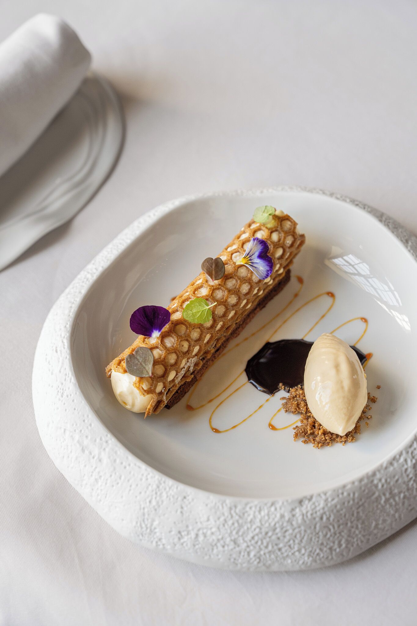 Un postre sobre un plato blanco con borde texturizado, un mantel de servilleta enrollado y un mantel blanco.