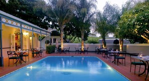 A poolside area with tables and chairs, a building with a patterned facade, and trees in the background. - Grande Roche Hotel (Paarl)