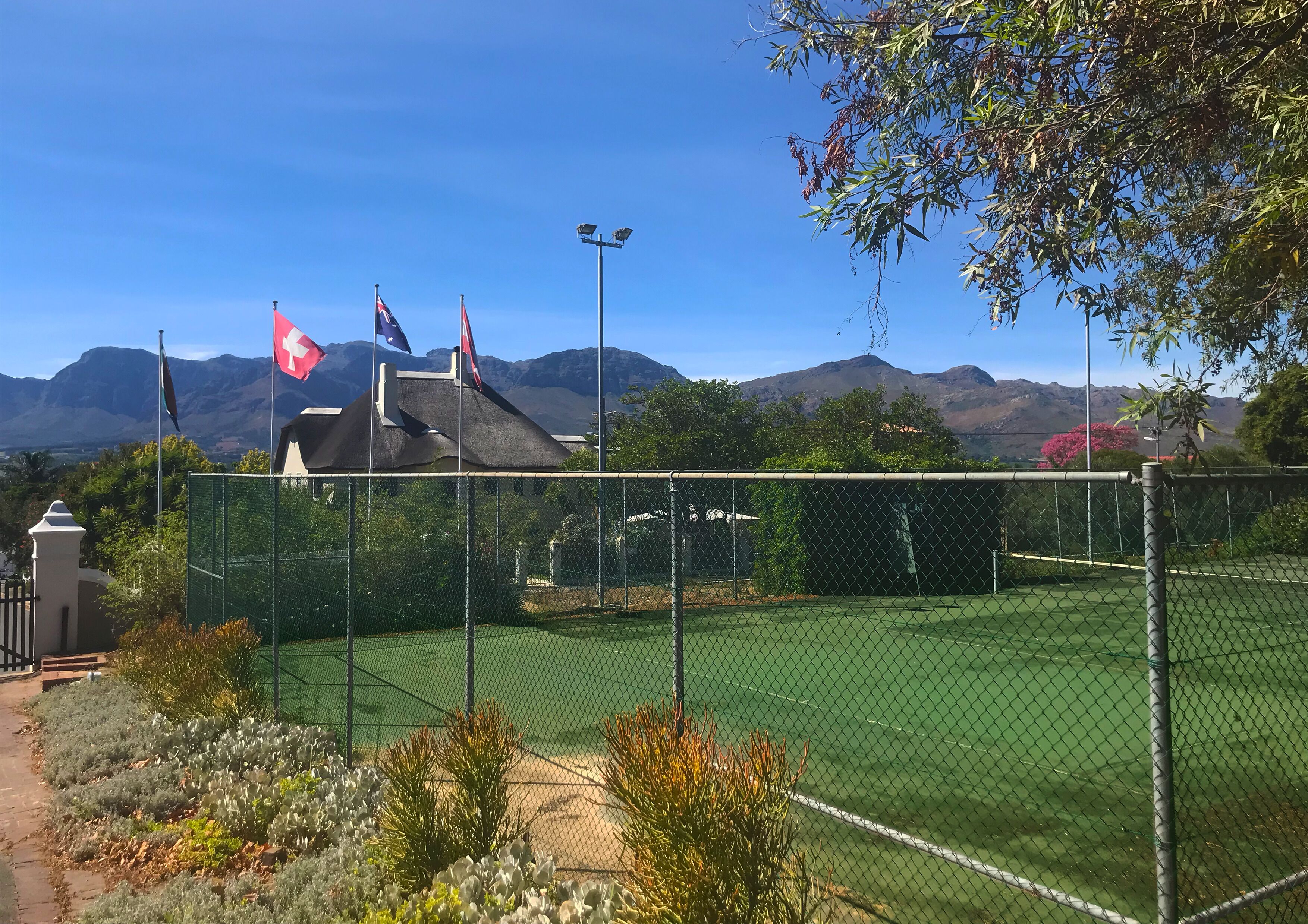 Een omheind tennisveld met vlaggen, omgeven door groen en bergen op de achtergrond.