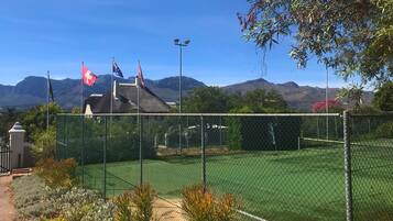 Een omheind tennisveld met vlaggen, omgeven door groen en bergen op de achtergrond.