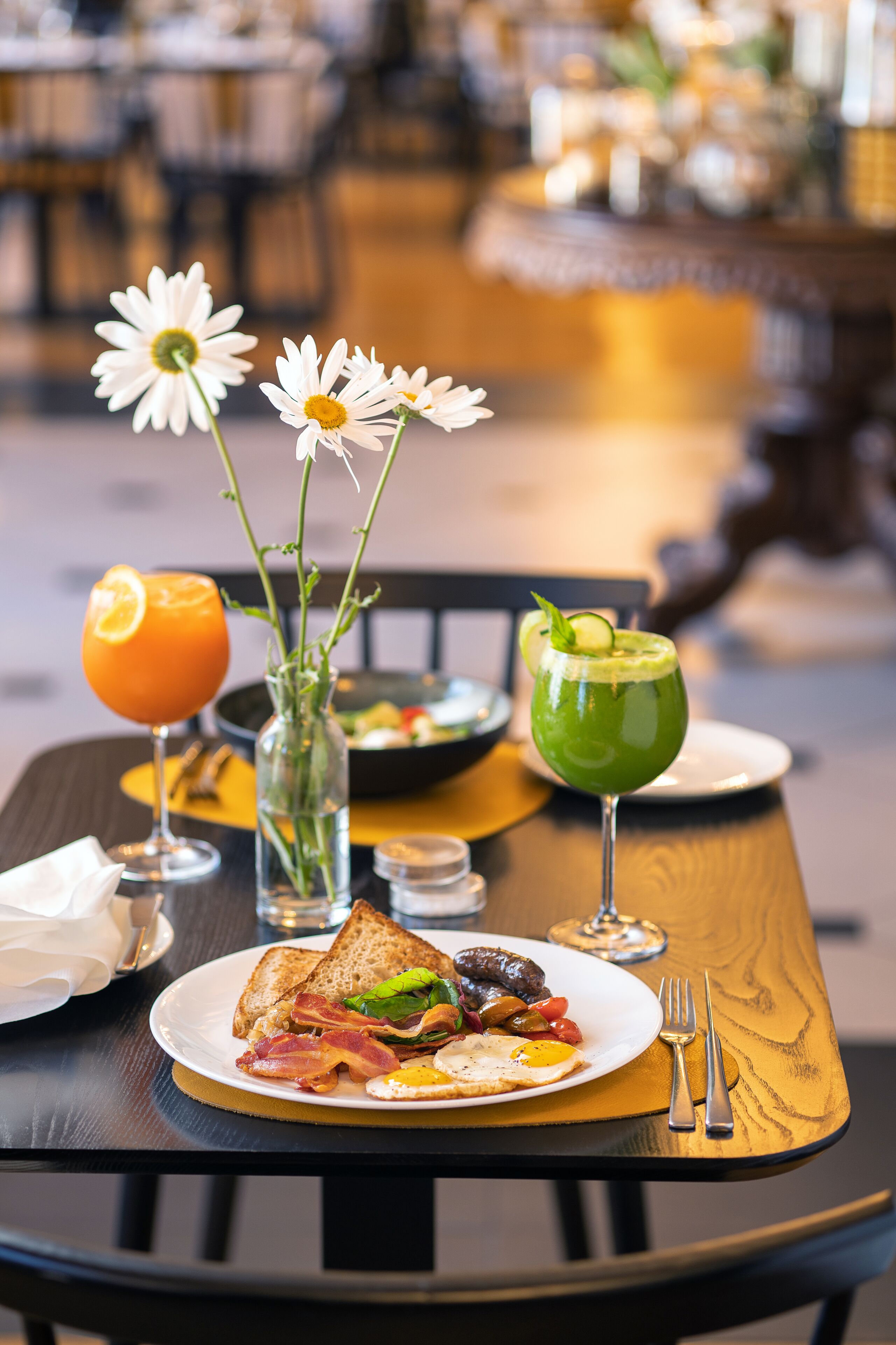 A breakfast table with a plate of eggs, bacon, toast, and a green drink. There are also two white flowers in a vase.
