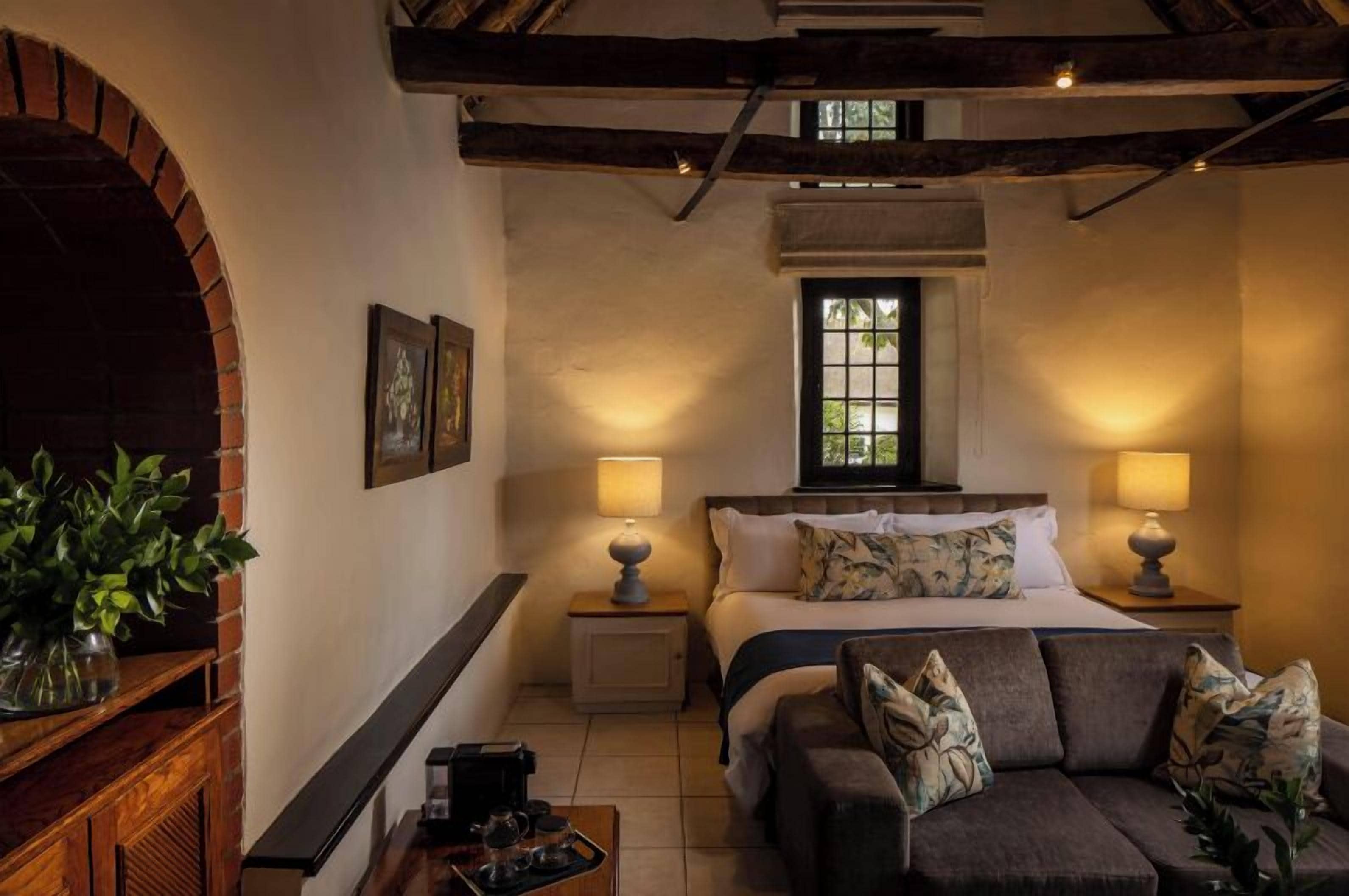 A cozy bedroom with a bed, two bedside lamps, a window, a wooden bench, and a plant.