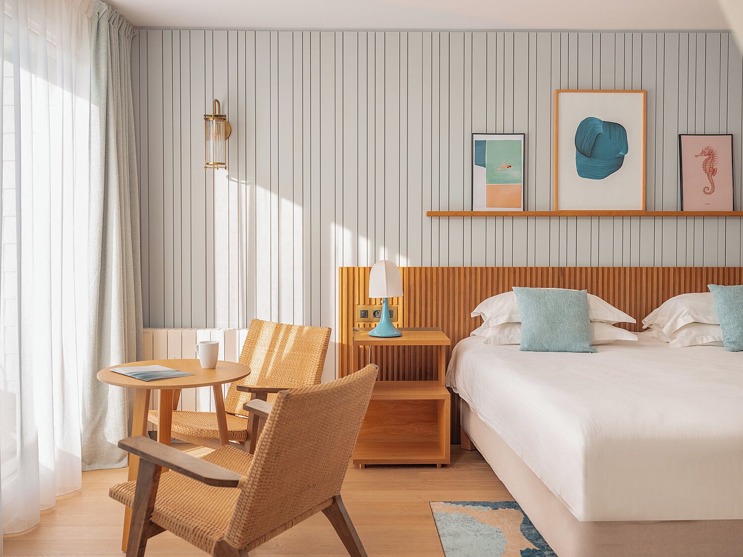 Habitación de hotel con cama, escritorio, silla y una mesita. Hay cortinas en la ventana, una lámpara en el escritorio y una obra de arte en la pared.