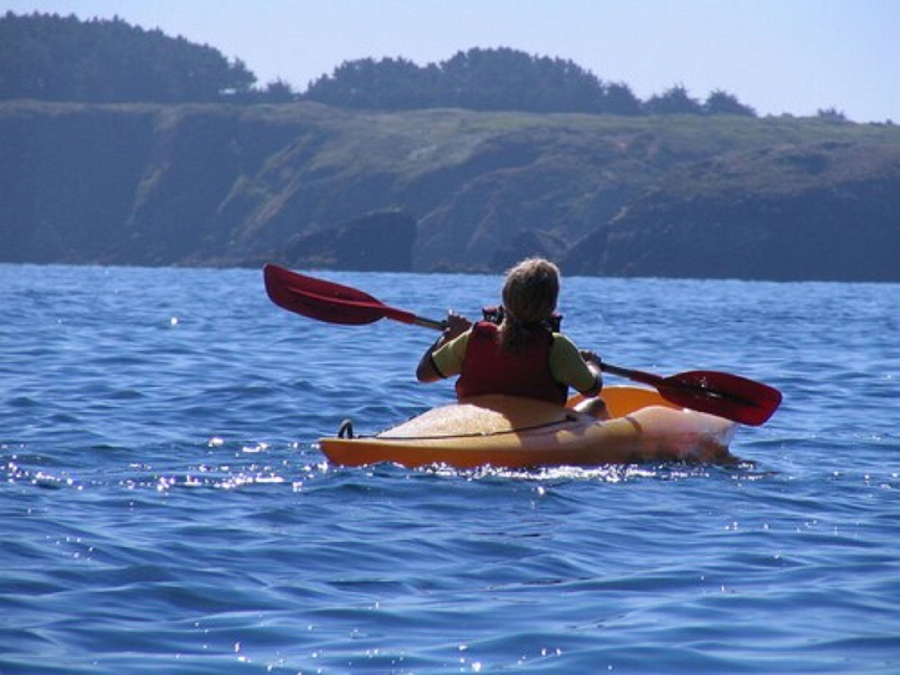 Eine Person paddelt mit einem Kajak auf dem Wasser, im Hintergrund eine hügelige Landschaft.