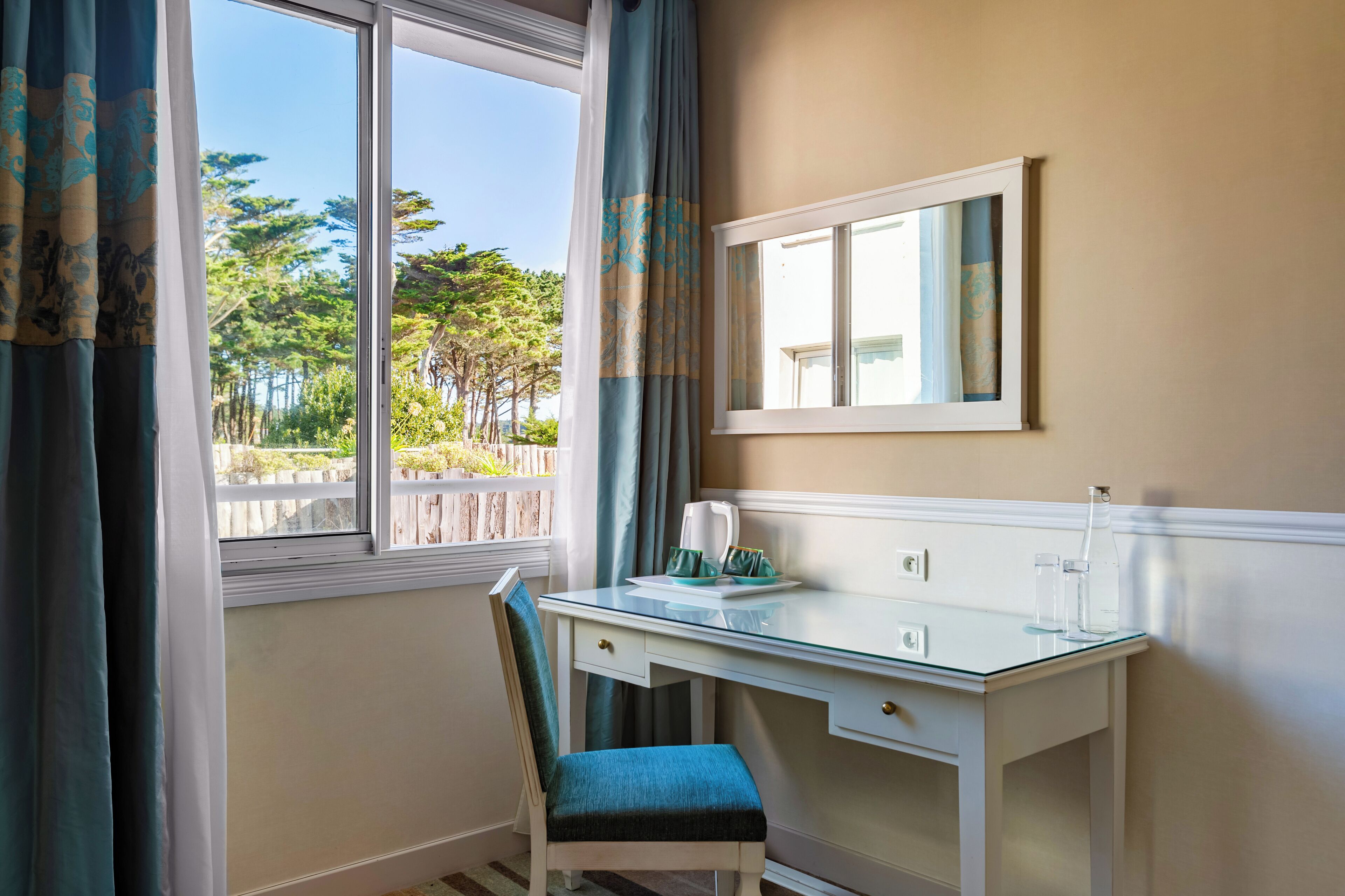 A room with a glass desk, a chair, a window with curtains, a mirror, and a view of trees and a fence.