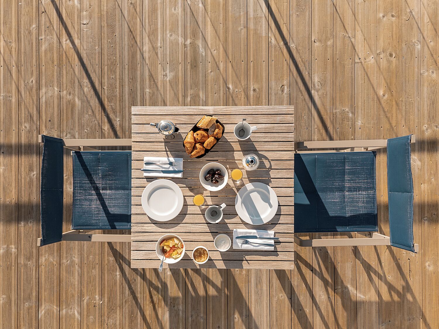 Una terraza de madera con una mesa dispuesta para el desayuno, que incluye cruasanes, fruta y café.