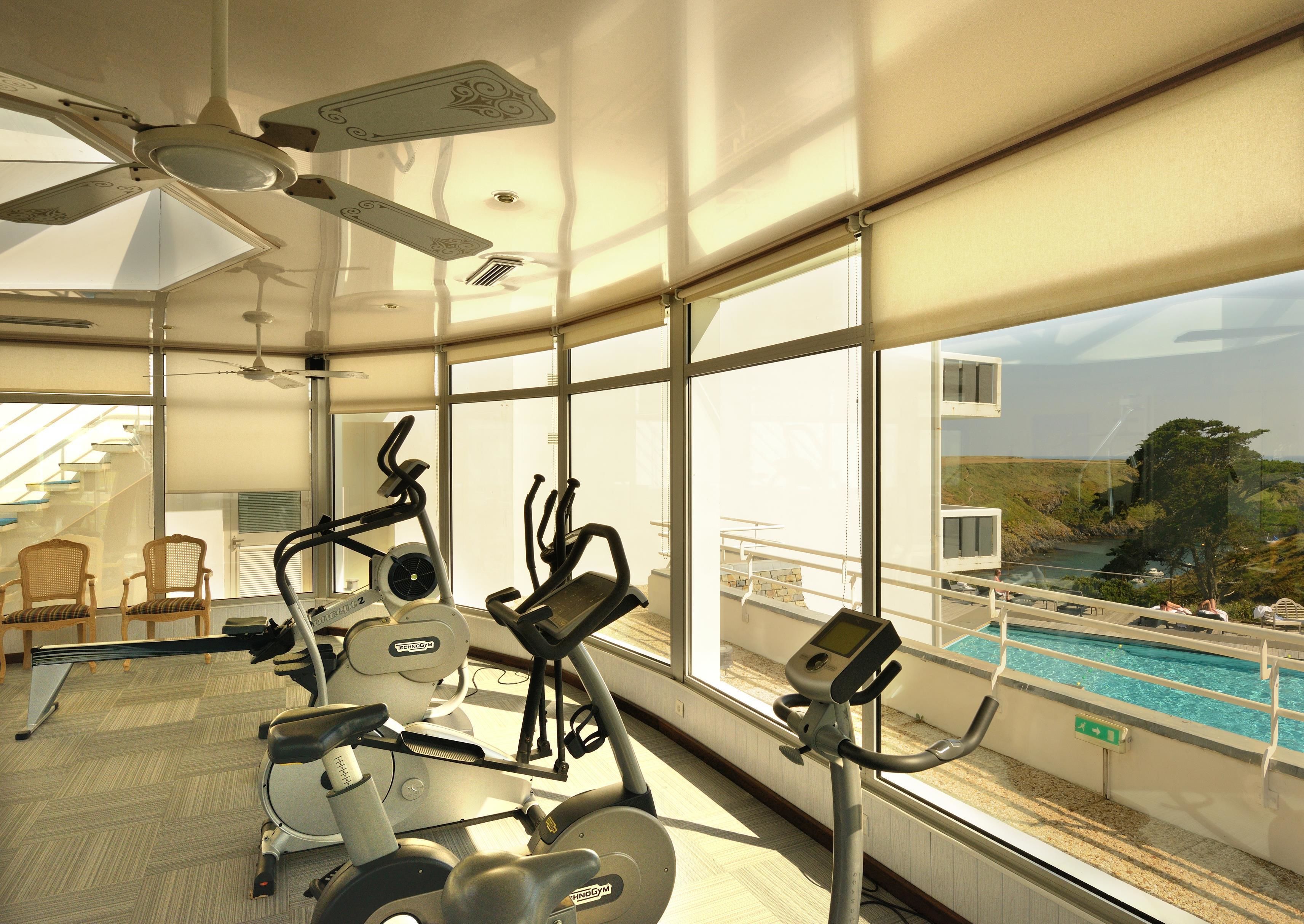 A gym with exercise equipment, large windows, and a view of a swimming pool.
