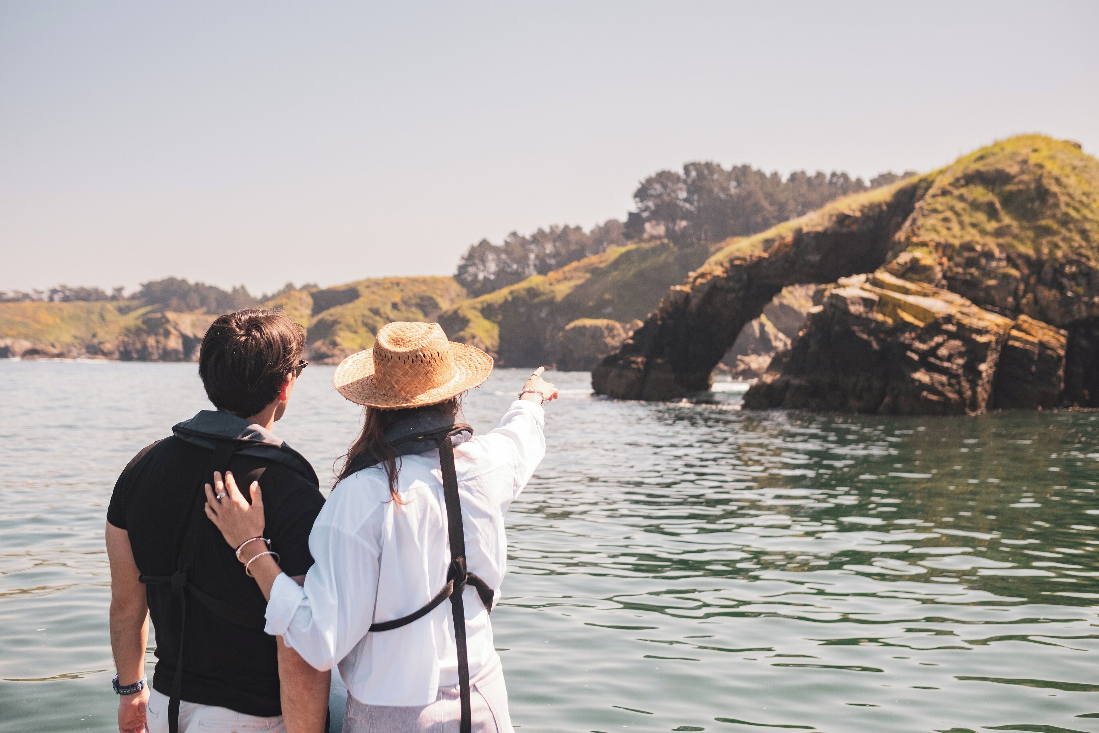 A couple standing by a body of water, pointing at a natural rock formation.
