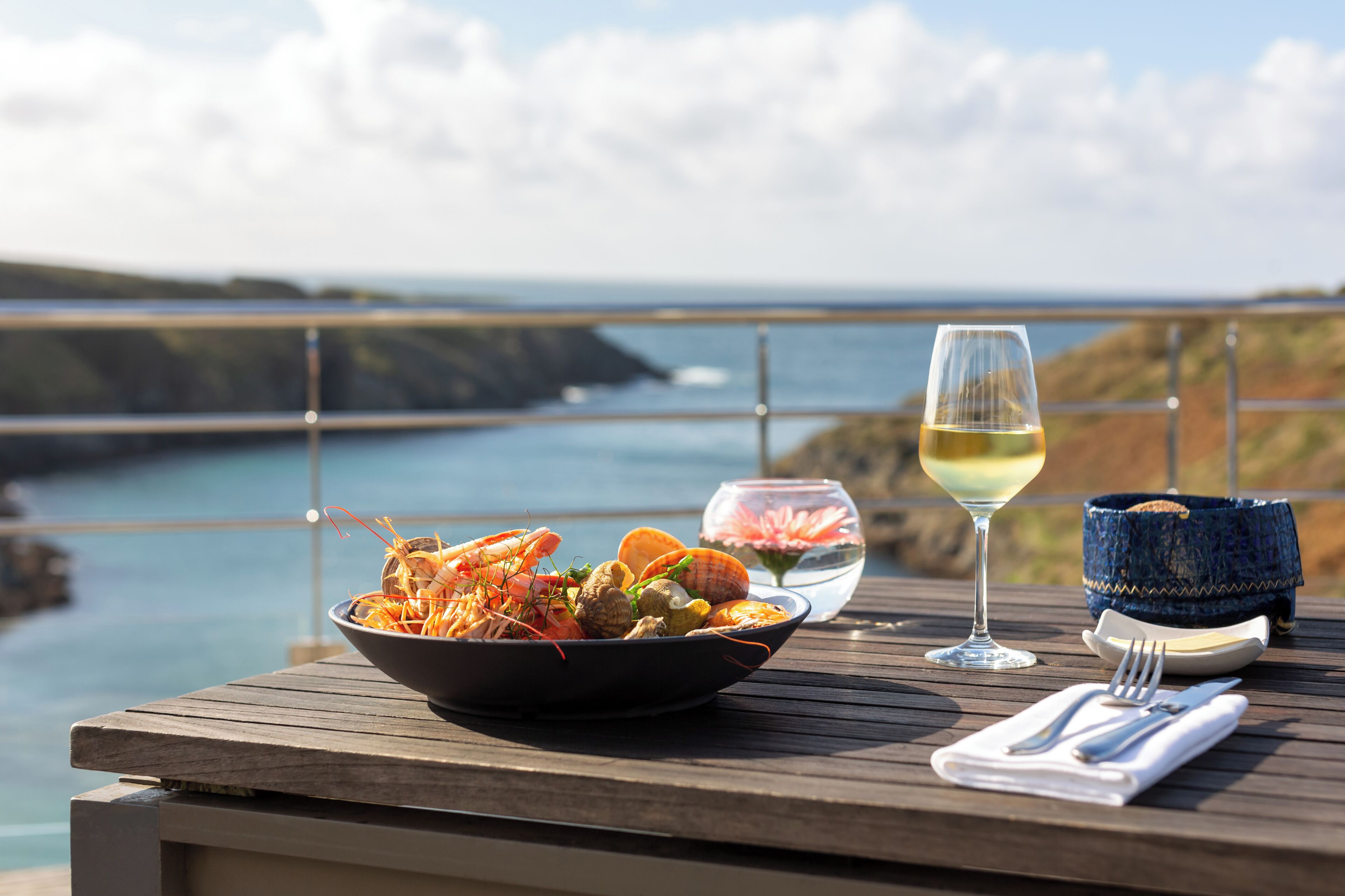 A dining table with seafood, a glass of white wine, and a basket of bread on a wooden deck overlooking a coastal landscape.