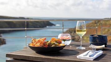 A dining table with seafood, a glass of white wine, and a basket of bread on a wooden deck overlooking a coastal landscape.