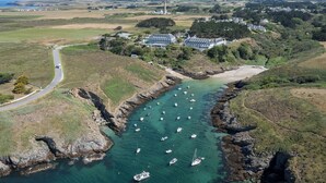 An aerial view of a coastal area with a large building surrounded by greenery, a narrow waterway with boats, and a road curving through the landscape. - Castel Clara Thalasso & Spa (Bangor)