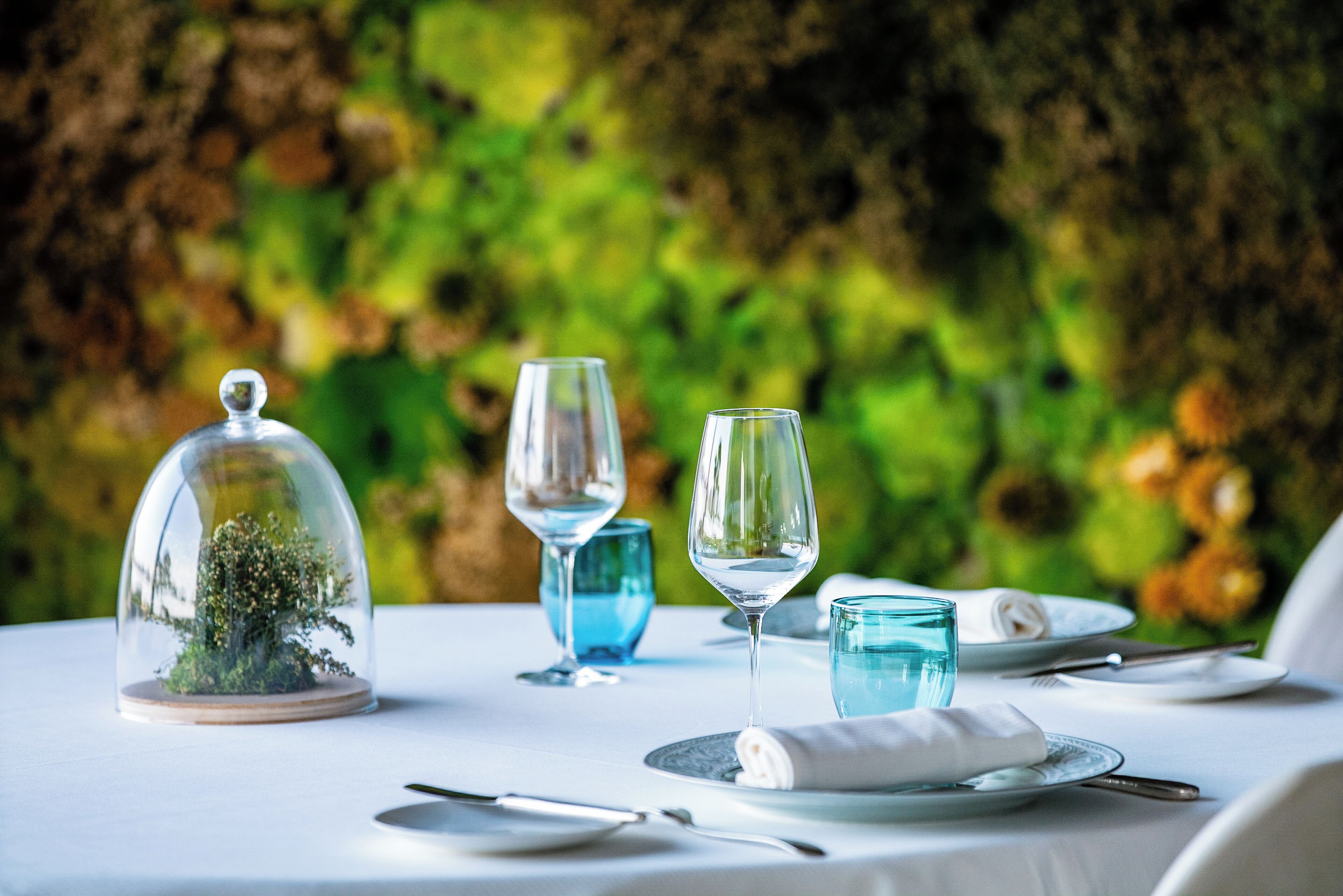 Une table dressée pour le dîner, ornée d’un centre de table en forme de cloche de verre, de verres à vin transparents et de verres à eau bleus.
