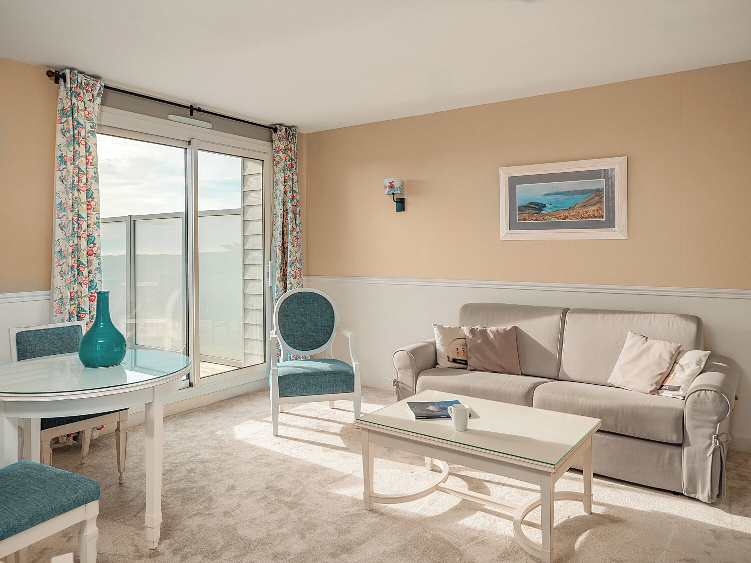 A living room with a glass table, a beige sofa, and a chair. There is a window with curtains, a framed picture on the wall, and a vase on the table.