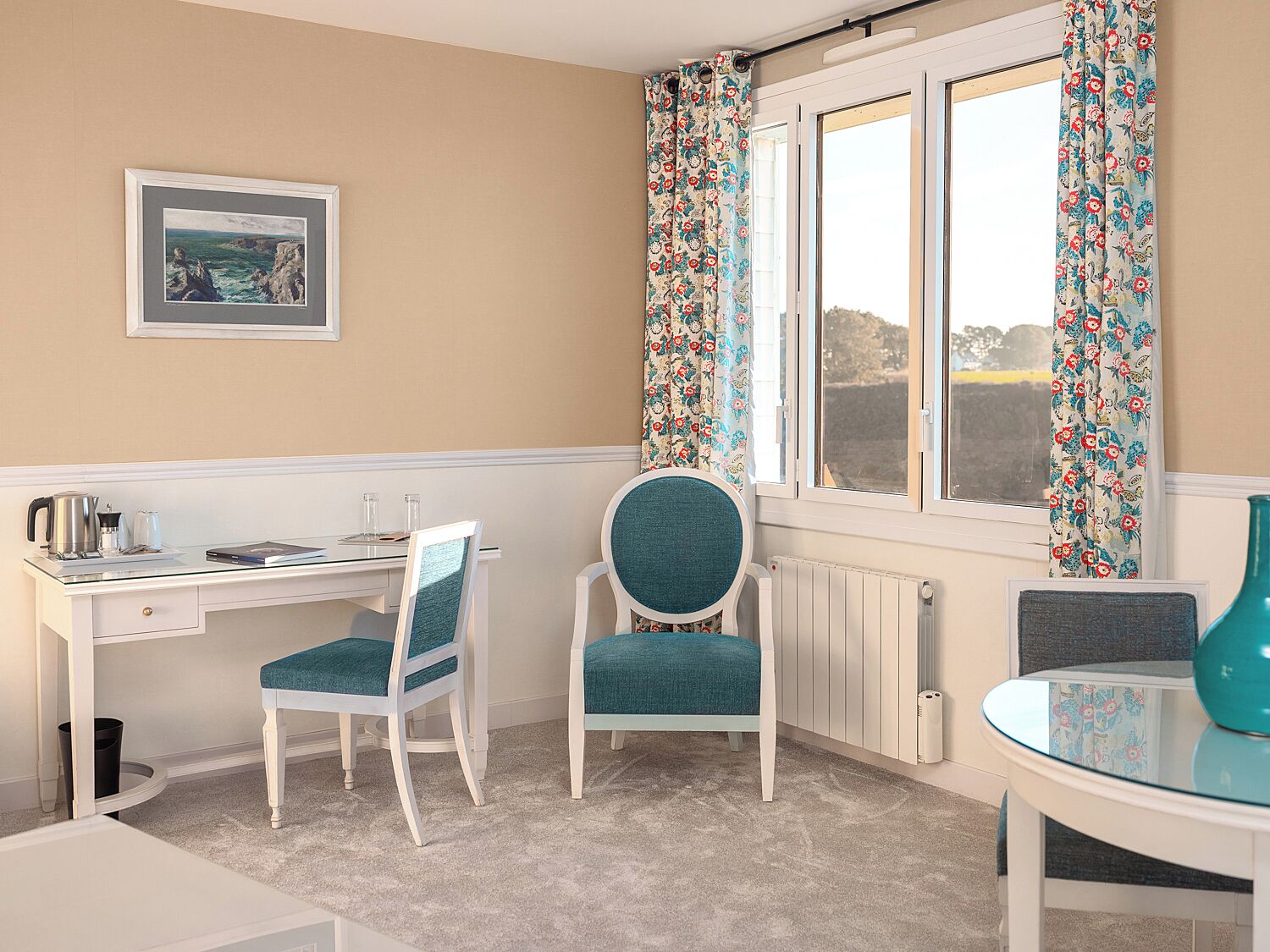 Une pièce avec un bureau blanc, deux chaises, une fenêtre ornée de rideaux à motifs floraux, un radiateur et une petite table sur laquelle est posé un vase.