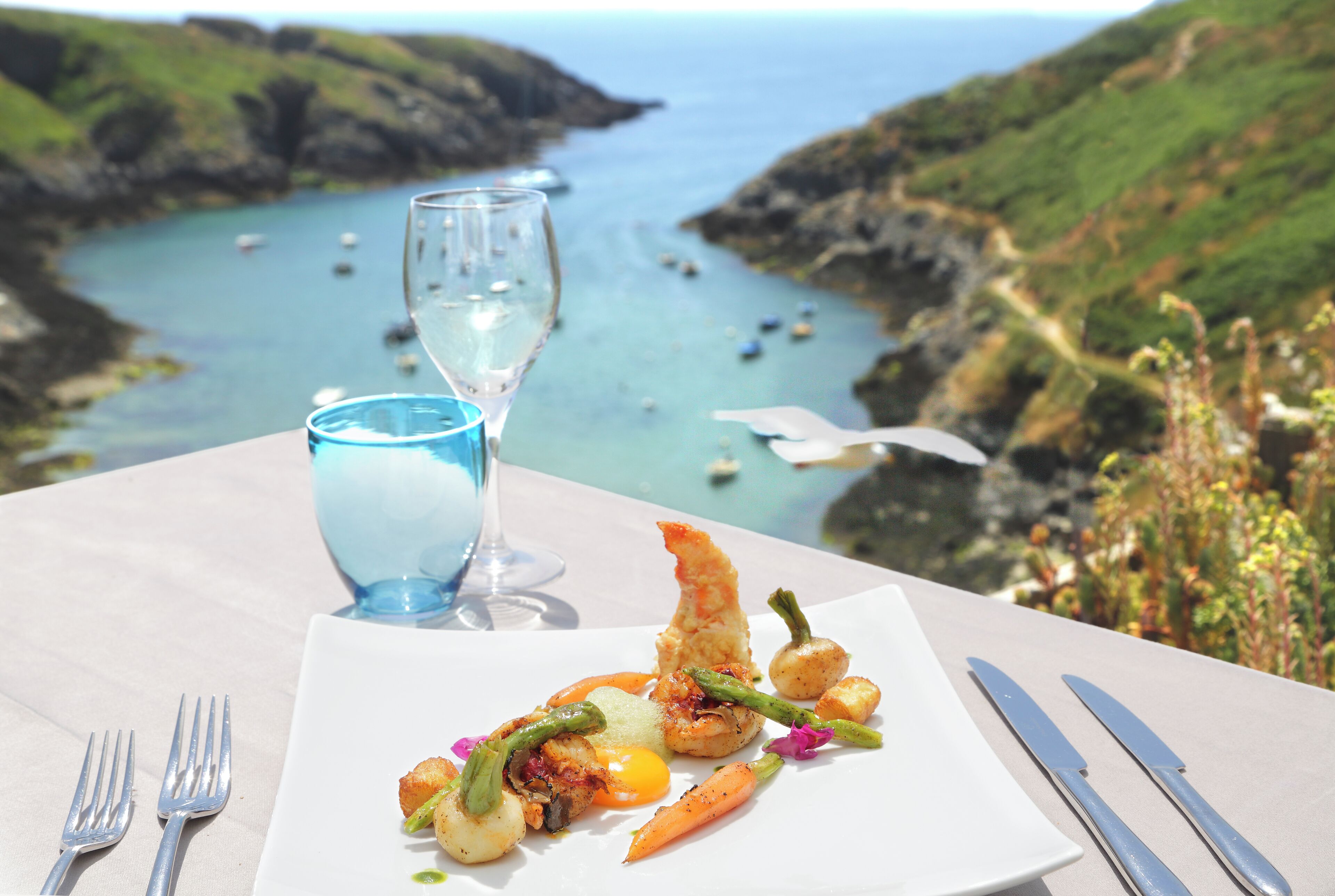 A table with a seafood dish, a glass of water, and cutlery. The background features a coastal view with boats and greenery.