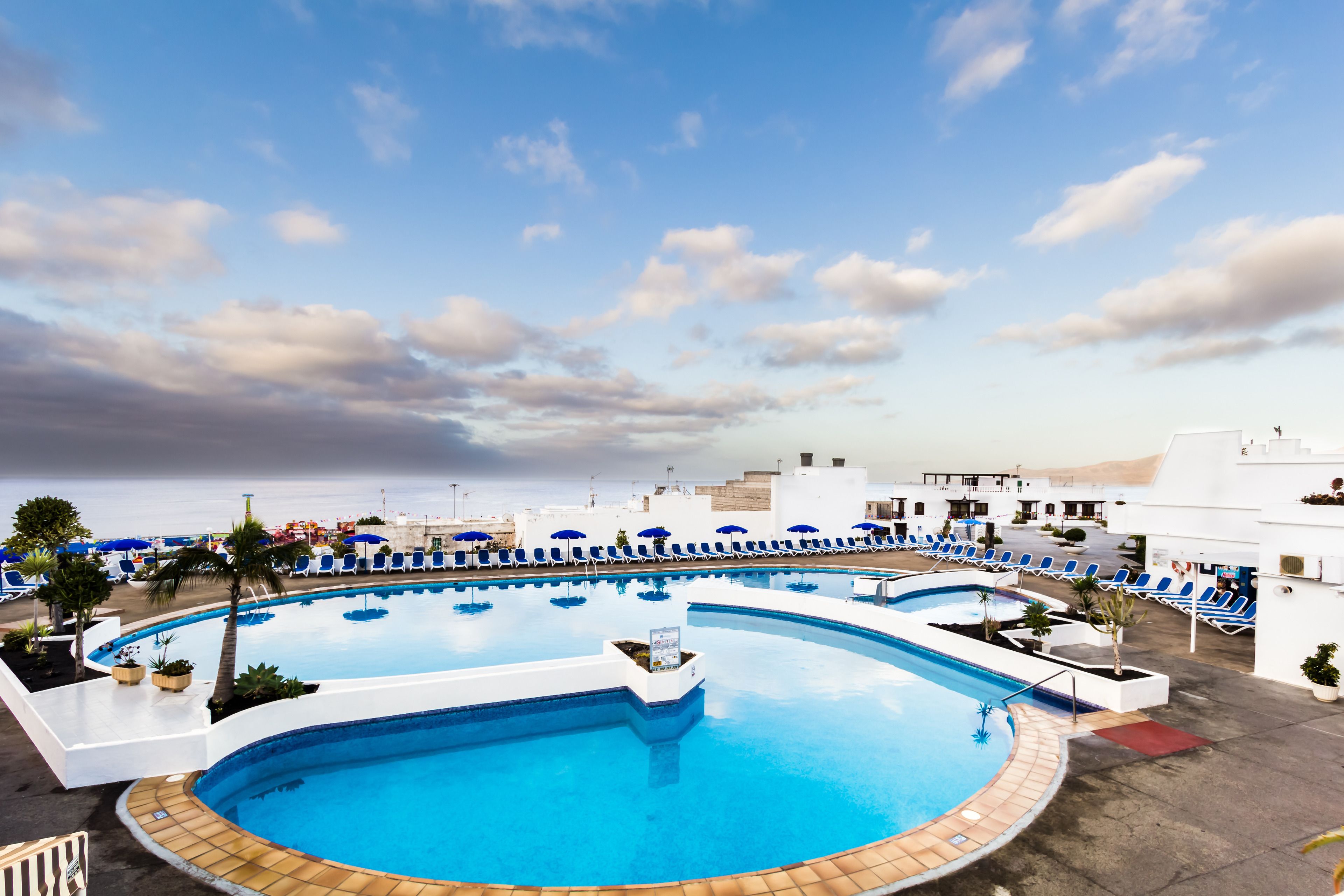 A large outdoor swimming pool with blue water, surrounded by white buildings and blue lounge chairs.
