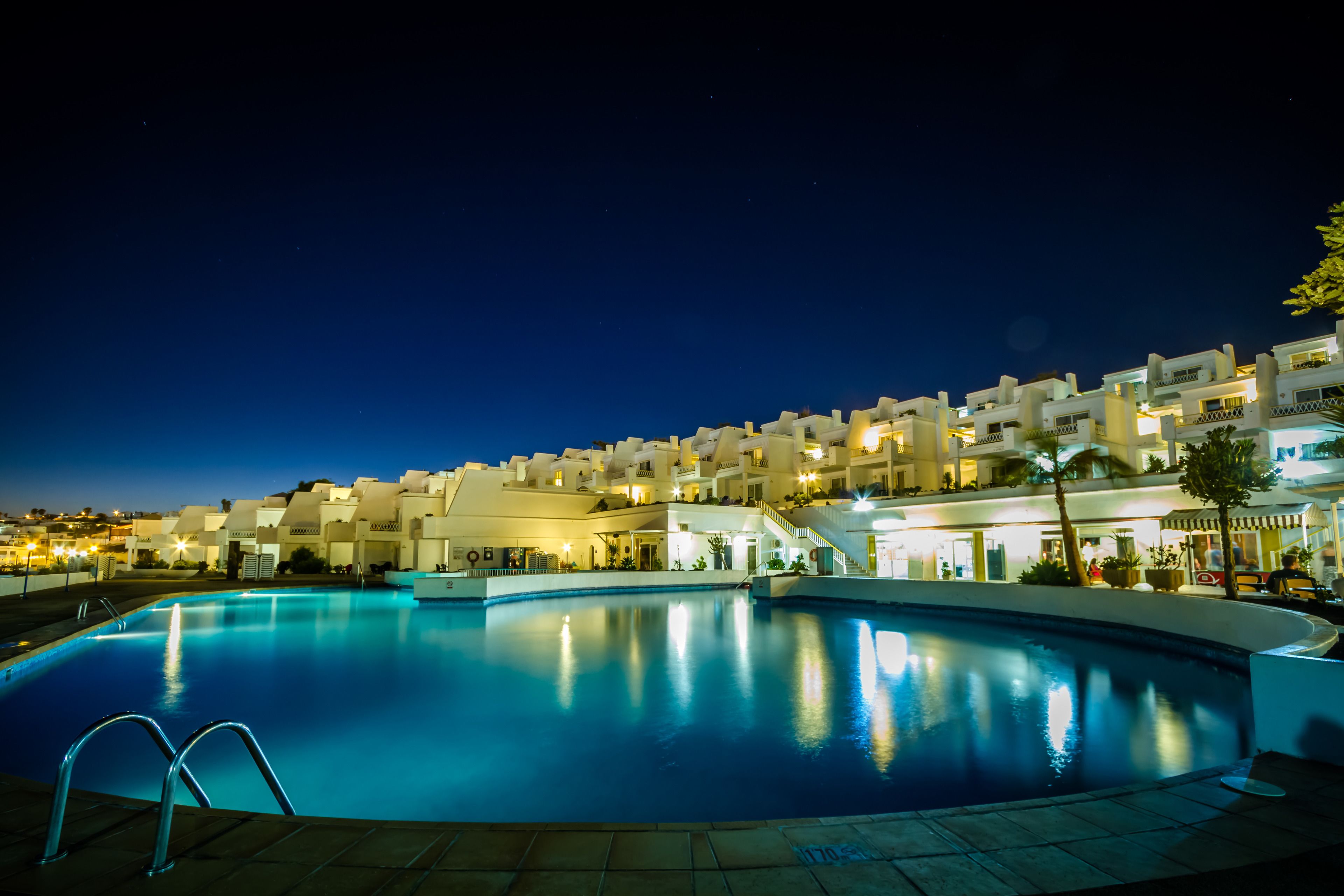 Un área de piscina iluminada de noche, con un edificio bien iluminado al fondo.