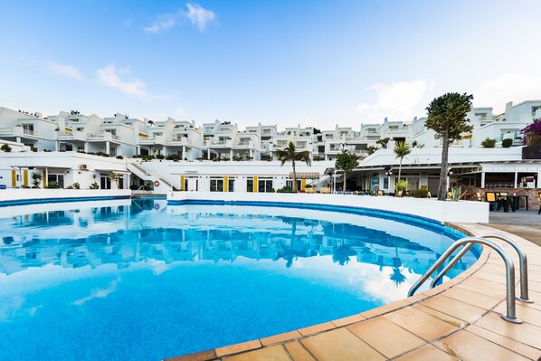 A swimming pool with a clear blue water, surrounded by white buildings with balconies and a restaurant area.