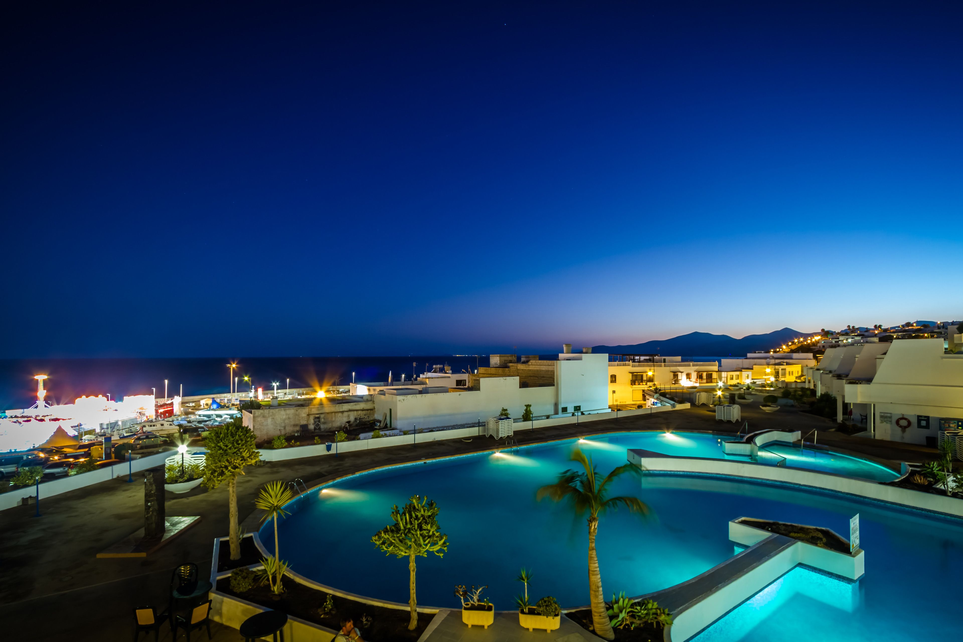 A nighttime view of a swimming pool area with a view of the sea and buildings in the background.