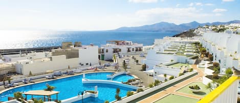 A resort with a large swimming pool, multiple tennis courts, and a view of the sea and mountains.