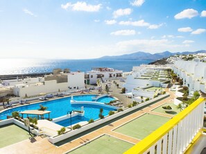 A resort with a large swimming pool, multiple tennis courts, and a view of the sea and mountains.