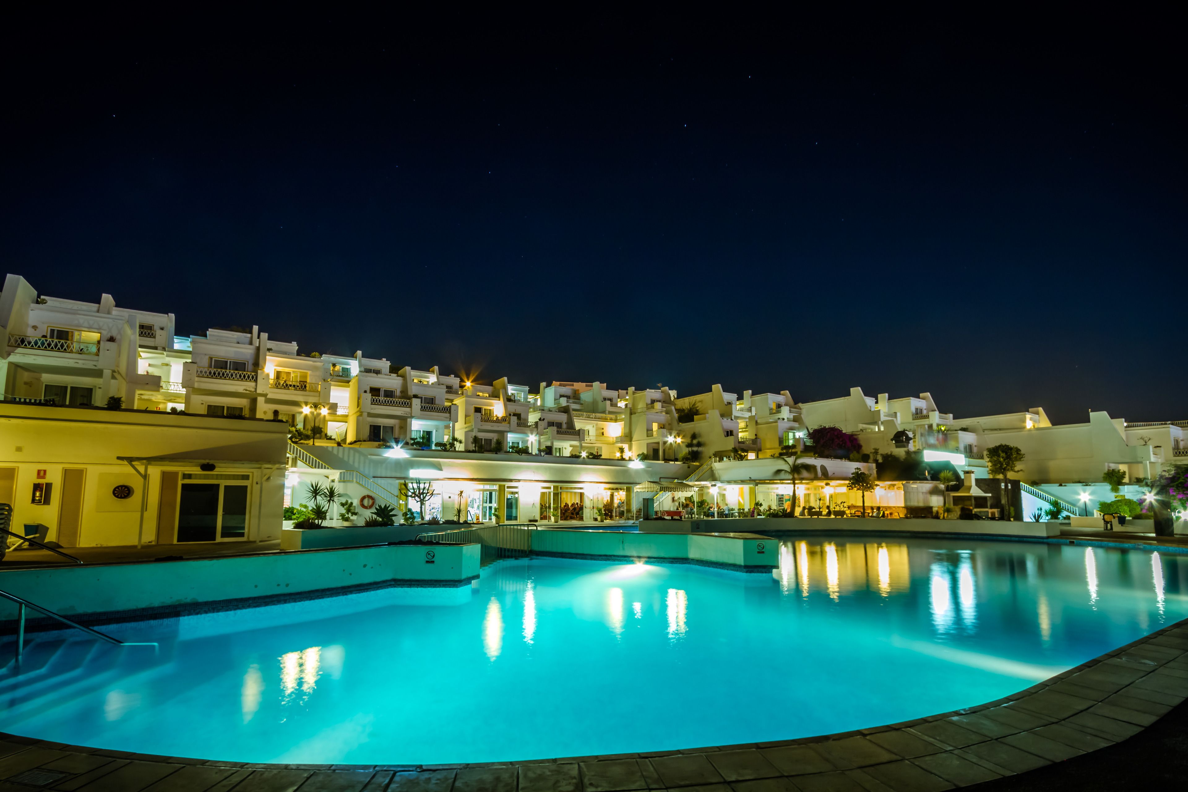 Vista nocturna de un hotel con piscina, balcones iluminados y una ladera llena de edificios.