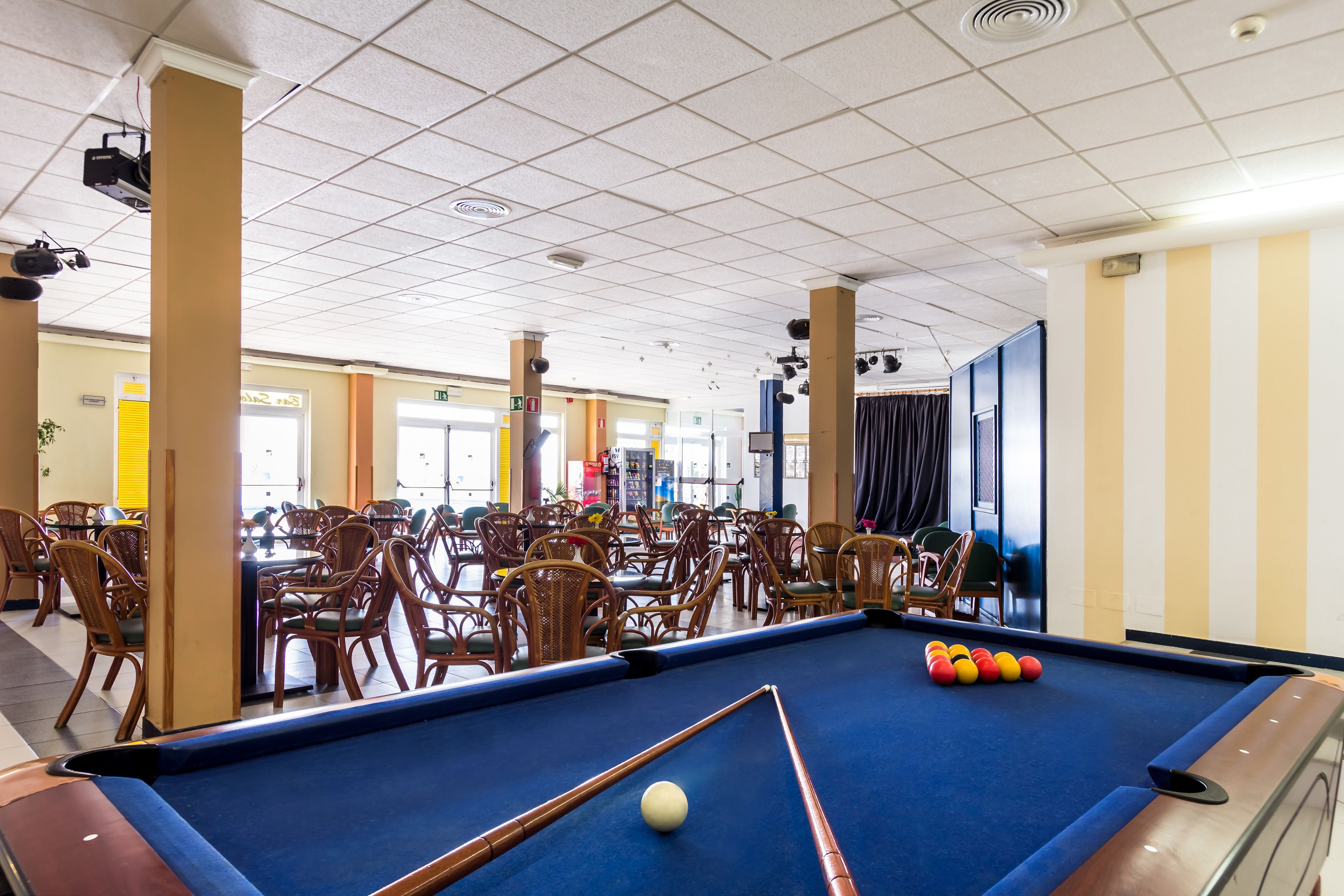 Ein Billardtisch mit einer weißen Spielball und bunten Kugeln, umgeben von Korbstühlen in einem Raum mit gefliestem Decken und gelb-weißen Streifenwänden.