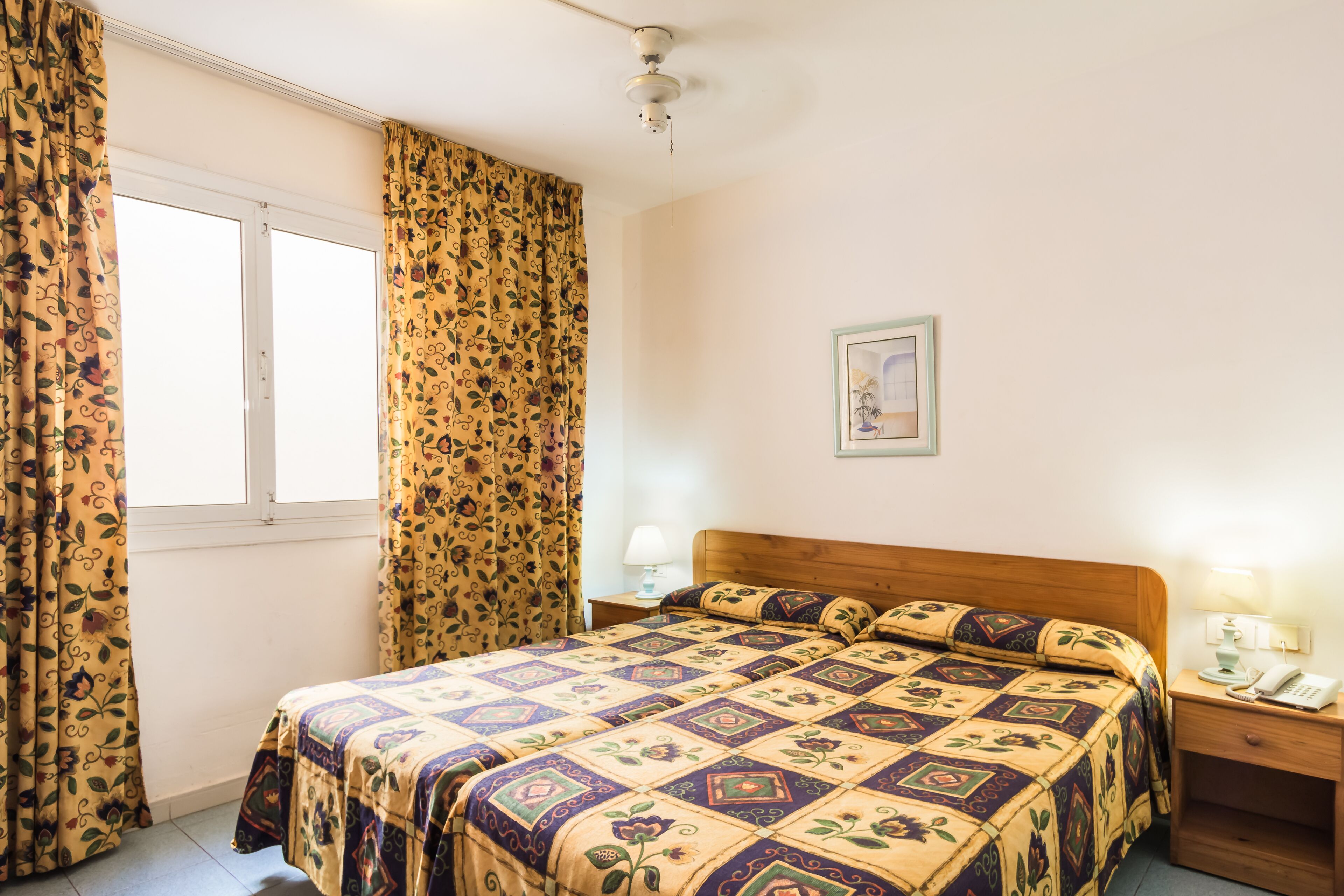 A bedroom with a bed, curtains, a bedside table with a lamp, a framed picture on the wall, and a ceiling fan.