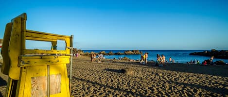 Una playa con un puesto de socorristas amarillo, formaciones rocosas en el agua y gente relajándose en la arena.