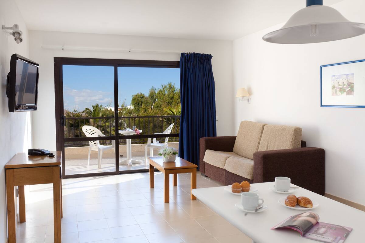 A hotel room with a balcony, a sofa, a table with pastries, a TV, and a framed picture on the wall.