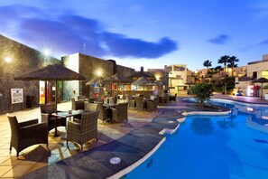 An outdoor pool area with lounge chairs and umbrellas, surrounded by buildings with palm trees. - BLUESEA Costa Teguise Gardens (Teguise)