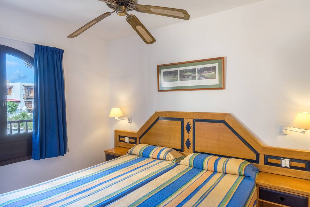 A hotel room with a bed, a ceiling fan, a window with curtains, and a framed picture on the wall.