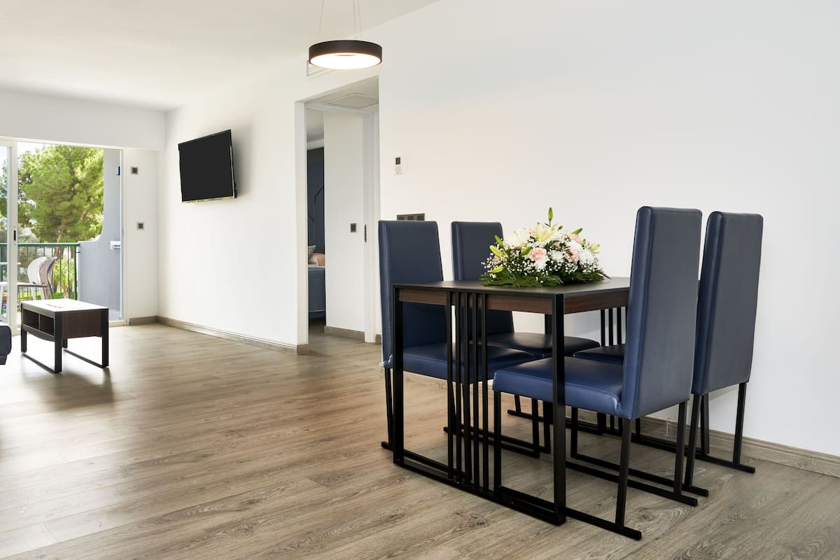 Un salon moderne comprenant un canapé, une table à manger avec des chaises, un téléviseur fixé au mur et un balcon offrant une vue sur les arbres.