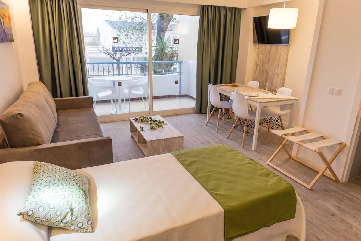 Un salon moderne comprenant un canapé, une petite table recouverte d’une nappe verte, une table à manger avec des chaises, et un téléviseur fixé au mur.
