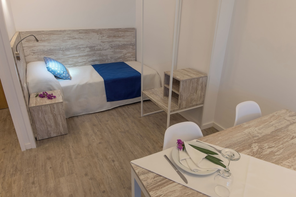 A bedroom with a bed, bedside table, and a small wooden nightstand. There is a dining table with a chair, a wine glass, and a plate.